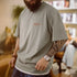 Man wearing a gray t-shirt with a logo, holding a patterned bag indoors.